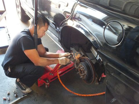 2003 Ford Crown Victoria - Kneeling worker grinds the exposed rear rotor of a black car, with wheel studs and hub clearly visible.
