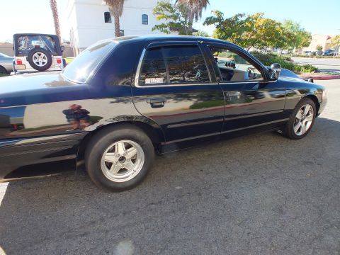 2003 Ford Crown Victoria - Sunlit passenger profile of a black sedan with a SuperMotors decal, Jeep parked behind.