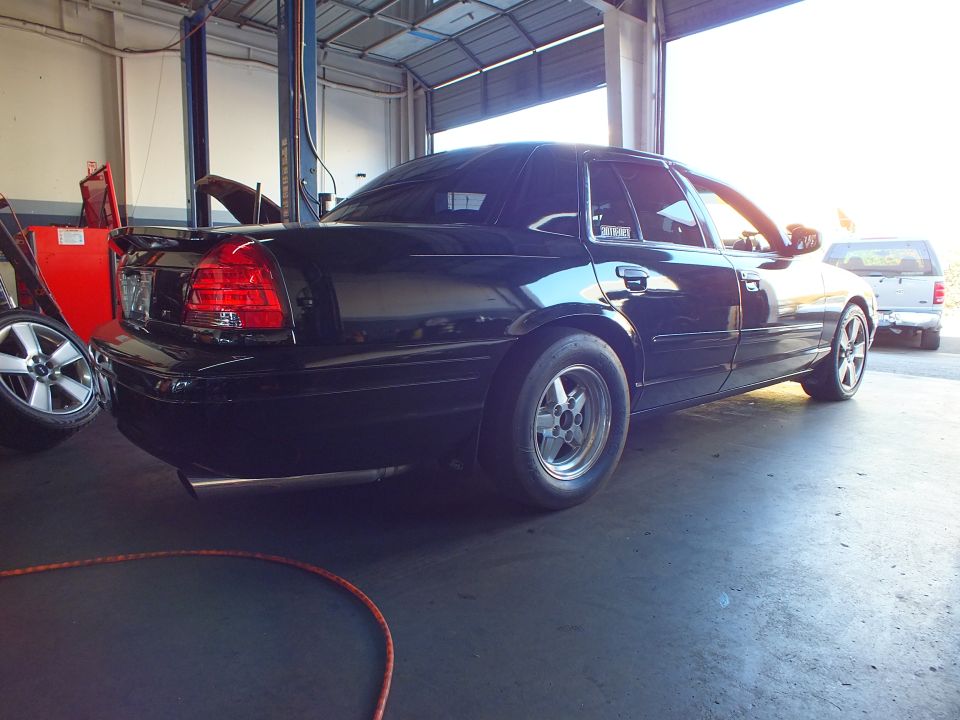 Black sedan rear three-quarter in shop bay with SUV and tire in background