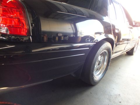 2003 Ford Crown Victoria - Low close-up of a wide rear tire and polished wheel under a black sedan in a shop.