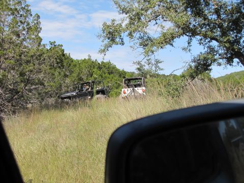 1995 Ford Bronco - Two off-road vehicles are seen driving through a grassy, wooded area on a sunny day.