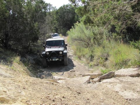 1995 Ford Bronco - Two off-road vehicles navigate a challenging dirt trail surrounded by trees and brush.