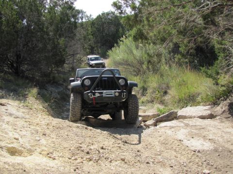1995 Ford Bronco - Two Jeeps navigate a challenging off-road path through a wooded area.