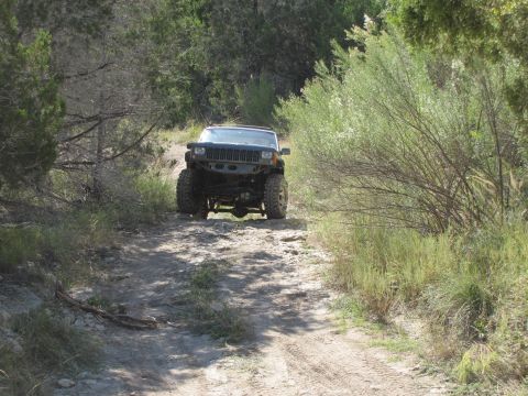 1995 Ford Bronco - A modified Jeep Cherokee XJ navigates a rough dirt trail surrounded by vegetation.