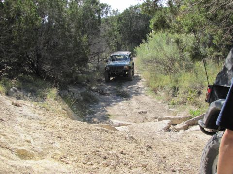 1995 Ford Bronco - Two off-road vehicles navigate a rough dirt path through a wooded area.