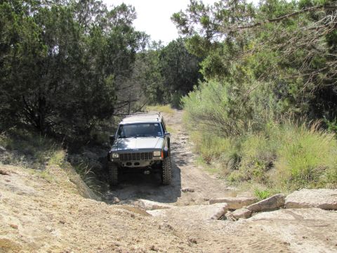 1995 Ford Bronco - A silver Jeep Cherokee XJ navigates a rocky, off-road trail surrounded by dense vegetation.