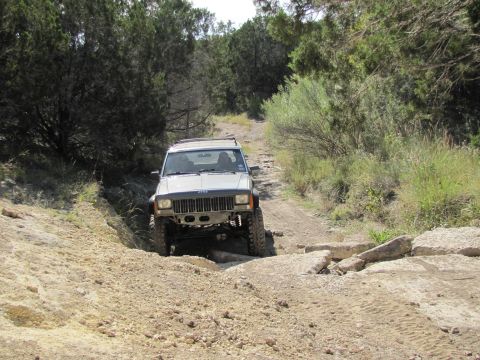 1995 Ford Bronco - White Jeep Cherokee XJ navigates a challenging rocky trail with dense foliage on either side.