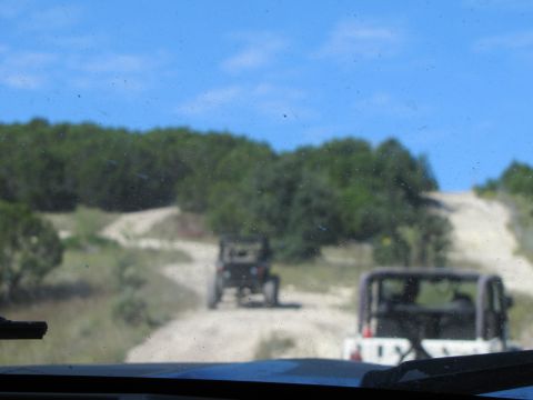 1995 Ford Bronco - Two off-road vehicles navigate a winding dirt path through a wooded area under a clear blue sky.