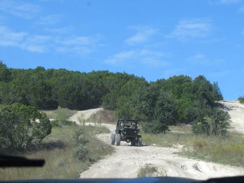 1995 Ford Bronco - An off-road vehicle navigates a dusty trail through a wooded landscape under a bright blue sky.