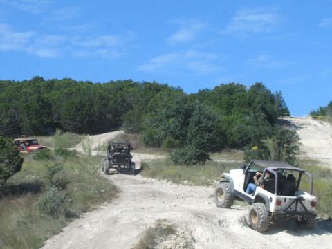 1995 Ford Bronco - Multiple off-road vehicles navigate a dusty trail surrounded by trees under a clear blue sky.