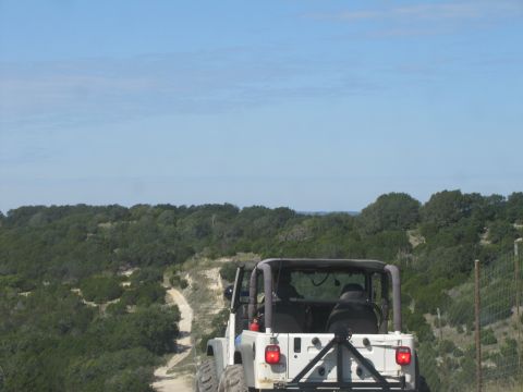 1995 Ford Bronco - A white Jeep Wrangler drives on a dirt road through a hilly, tree-covered landscape under a clear blue sky.
