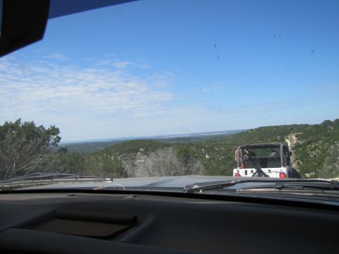 1995 Ford Bronco - Two vehicles travel on a dirt road overlooking a vast, tree-covered landscape under a partly cloudy sky.