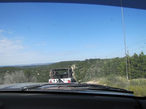1995 Ford Bronco - Two off-road vehicles navigate a dusty trail through a wooded, hilly landscape under a clear blue sky.