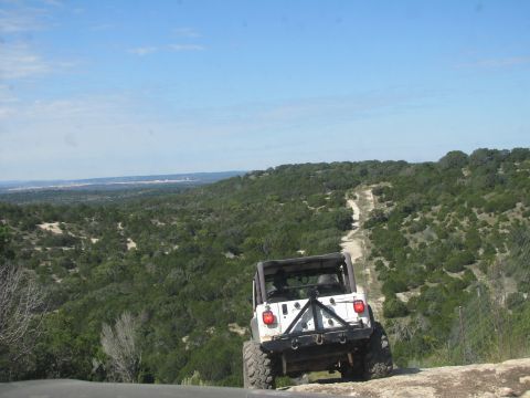 1995 Ford Bronco - A white Jeep Wrangler navigates a winding dirt road through a wooded, hilly landscape under a clear blue sky.