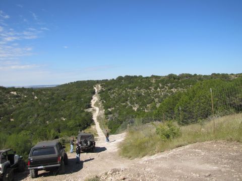 1995 Ford Bronco - Multiple off-road vehicles navigate a dusty trail winding through a dense, green landscape on a sunny day.