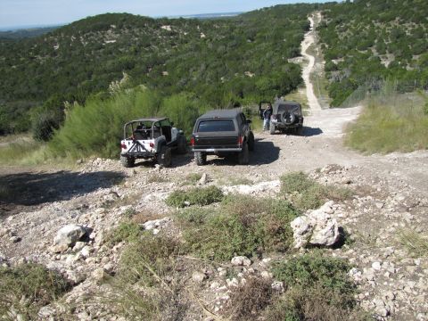 1995 Ford Bronco - Three off-road vehicles navigate a steep, rocky trail in a hilly, wooded area.