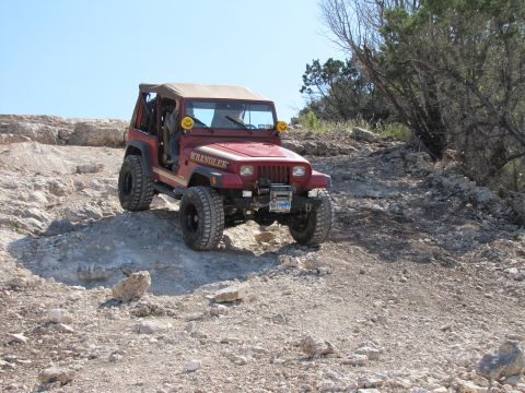 1995 Ford Bronco - A red Jeep Wrangler navigates a challenging rocky terrain under a clear blue sky.