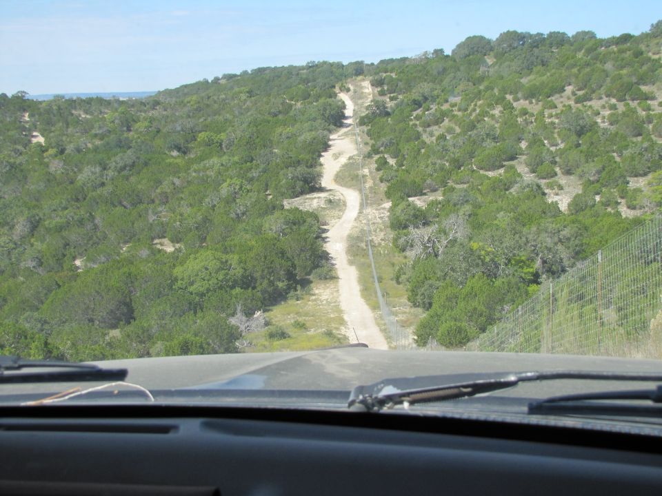 Interior view from a vehicle driving on a winding dirt road through a green, hilly landscape