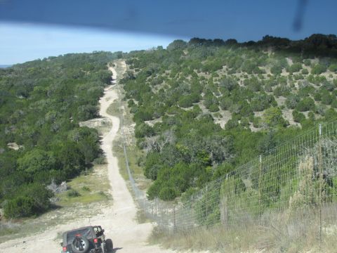 1995 Ford Bronco - A Jeep Wrangler navigates a dusty, off-road path surrounded by dense vegetation and a wire fence.