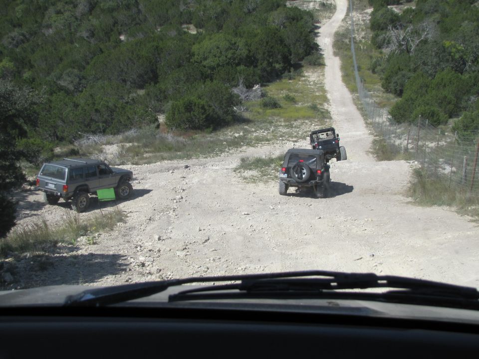 Three off-road vehicles on a dirt road in a wooded area. One SUV is on the left, two Jeeps are further up the road