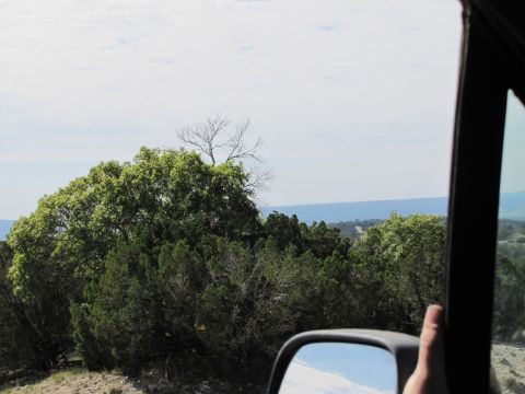 1995 Ford Bronco - View from a vehicle's side mirror shows a scenic landscape with lush green trees and a hazy horizon under a cloudy sky.