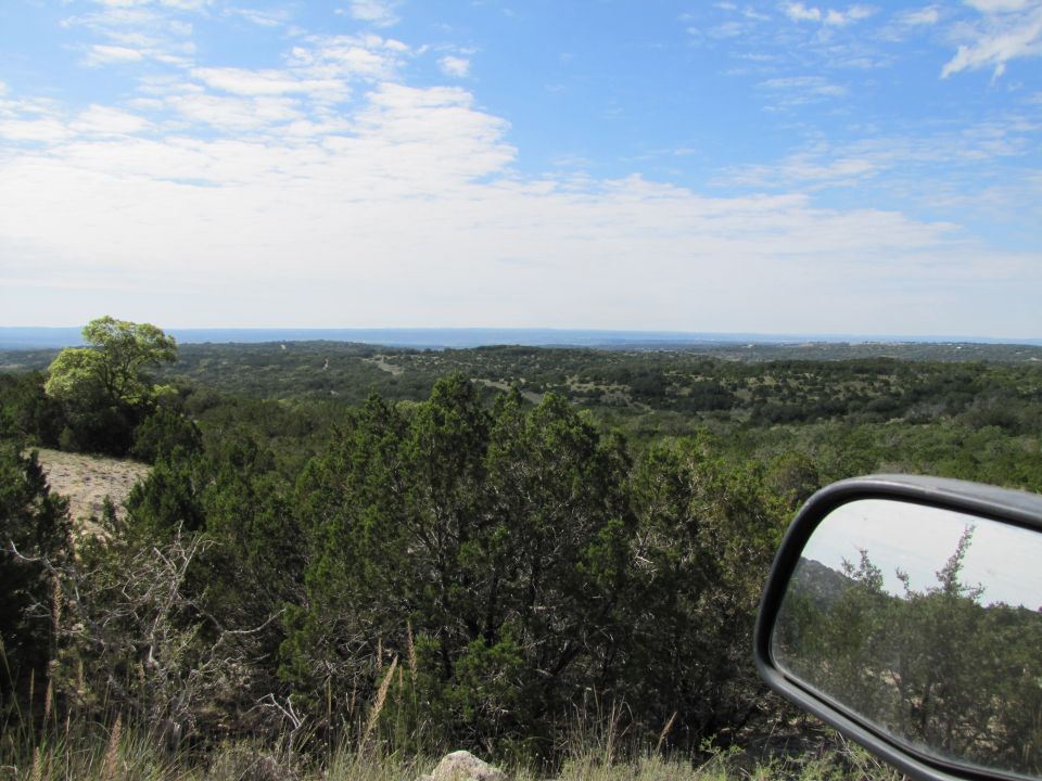 Side mirror reflecting a hilly, tree-covered landscape under a blue sky with clouds