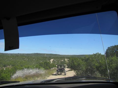 1995 Ford Bronco - Two off-road vehicles navigate a dusty trail through a dense, green, hilly terrain under a clear blue sky.
