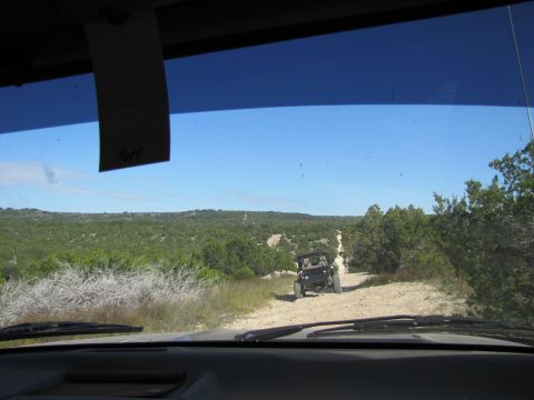 1995 Ford Bronco - Two vehicles driving on a dirt road through a wooded, hilly landscape under a clear blue sky.