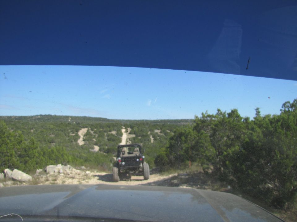 View from inside a vehicle looking at another off-road vehicle on a dirt path in a wooded, hilly area