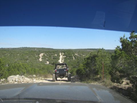 1995 Ford Bronco - Two off-road vehicles navigate a winding dirt path through a dense, green landscape under a clear blue sky.