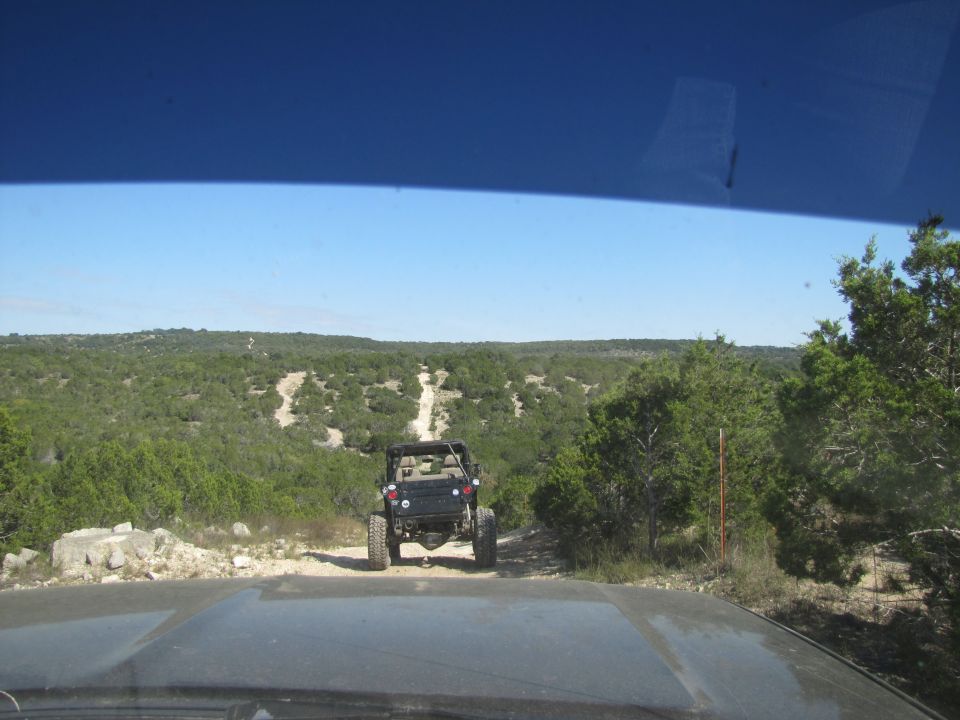 Jeep-like vehicle driving on a dirt trail through a wooded, hilly area. Another vehicle is behind it