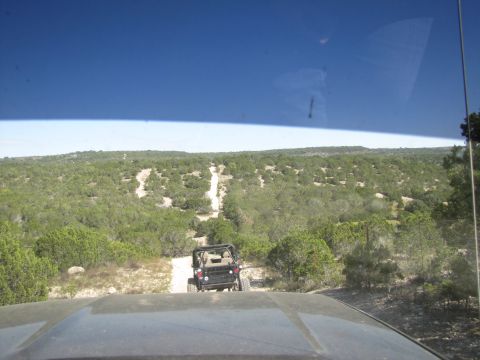 1995 Ford Bronco - Two off-road vehicles navigate a winding dirt path through a dense, green landscape under a clear blue sky.