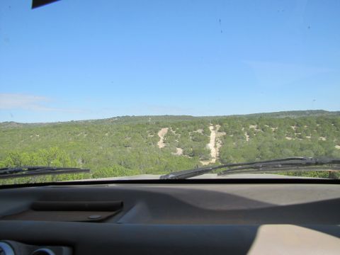 1995 Ford Bronco - View from inside a vehicle shows a dirt road ahead, leading through a landscape of green trees and hills under a bright blue sky.