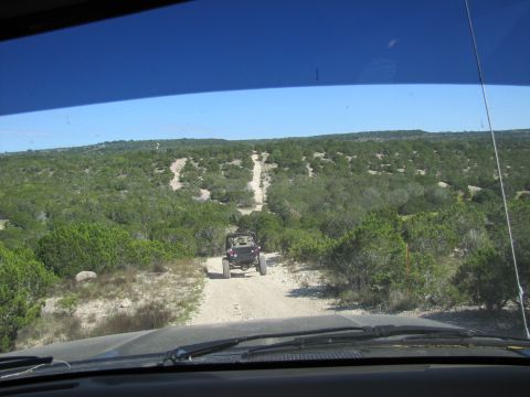 1995 Ford Bronco - Two off-road vehicles navigate a dusty trail winding through a dense, green, and hilly terrain under a clear blue sky.