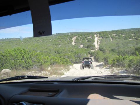 1995 Ford Bronco - Two off-road vehicles navigate a challenging dirt path through a , tree-covered landscape under a clear blue sky.