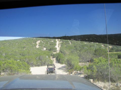 1995 Ford Bronco - Two off-road vehicles navigate a dusty trail amidst dense green foliage under a clear blue sky.