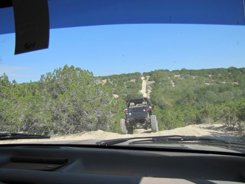 1995 Ford Bronco - A vehicle navigates a steep, dusty trail through a landscape dotted with scrubby trees under a clear blue sky.