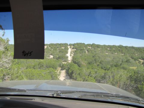 1995 Ford Bronco - Driving on a dirt road through a scenic, wooded area with hills under a clear blue sky.