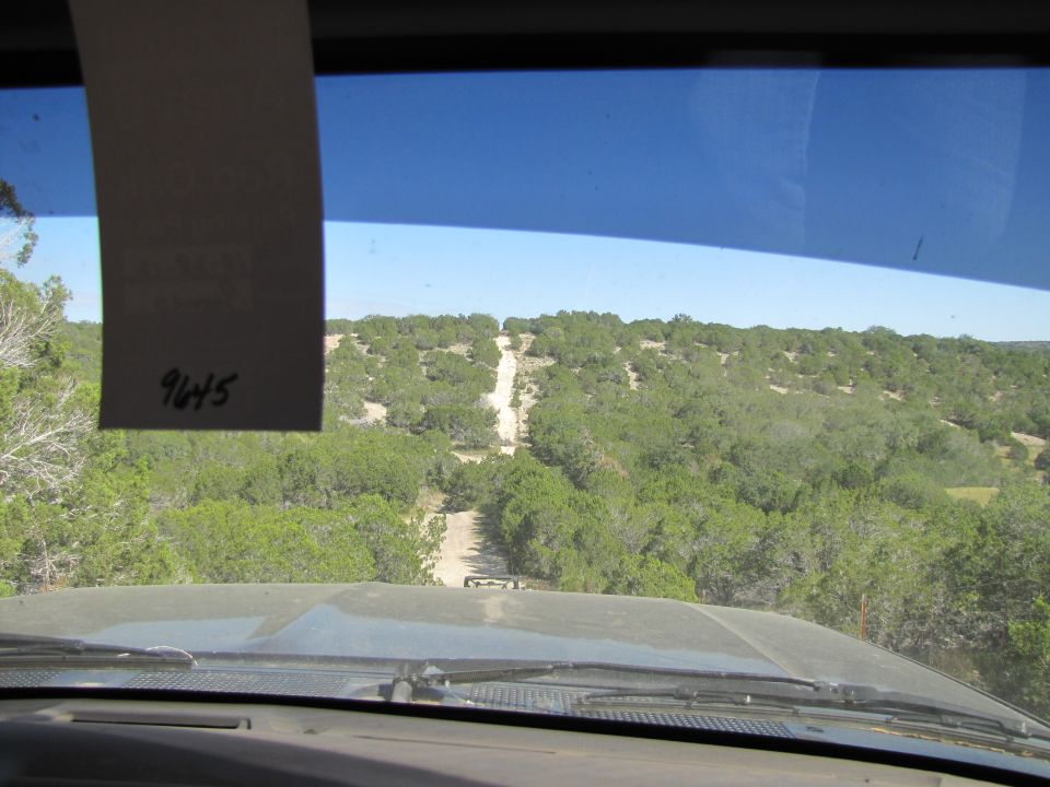 View from inside a vehicle looking at a dirt road winding through a hilly, tree-covered landscape. A tag with '9645' is visible on the windshield