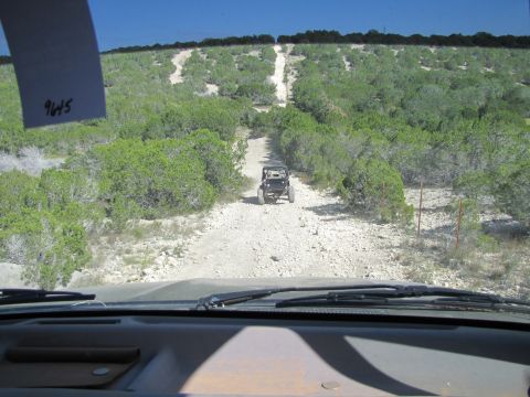 1995 Ford Bronco - Two vehicles navigate a rough, unpaved road surrounded by dense vegetation under a clear blue sky.