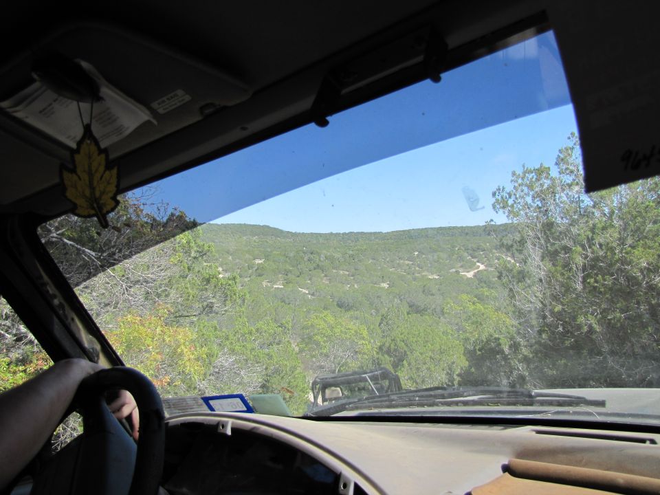 Interior view of a vehicle driving on a dirt road towards a scenic landscape. Another vehicle is visible ahead
