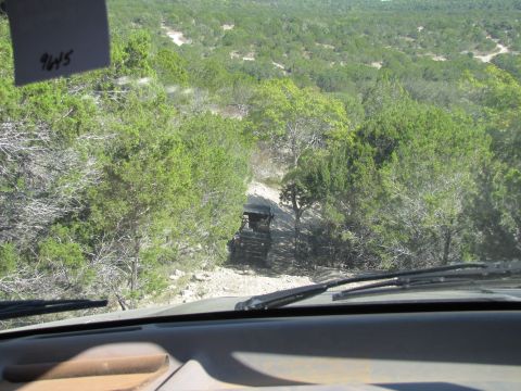 1995 Ford Bronco - View from inside a vehicle as a black SUV navigates a rough dirt road through a wooded area.