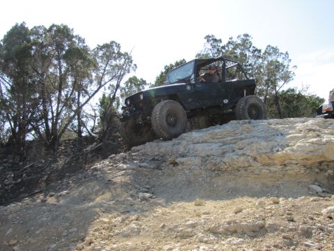 1995 Ford Bronco - A black Jeep Wrangler navigates a steep, rocky trail with trees in the background and another vehicle partially visible to the right.