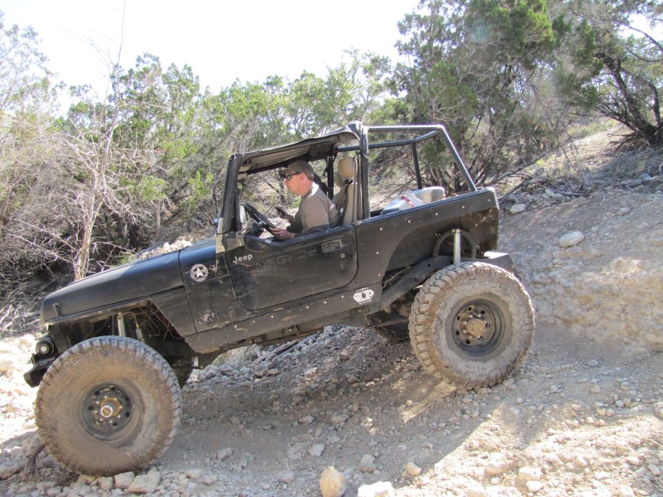 Black Jeep Wrangler TJ driving on a dirt and rock trail with trees in the background. Driver is wearing sunglasses