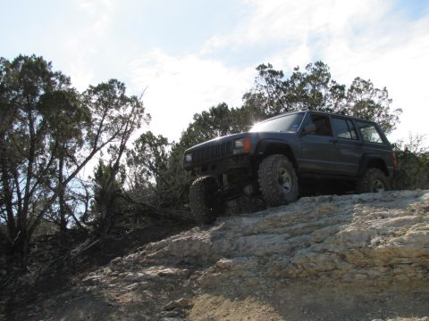 1995 Ford Bronco - A dark gray Jeep Cherokee XJ navigates a challenging rocky terrain under a bright sky.