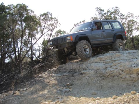 1995 Ford Bronco - A dark gray Jeep Cherokee is shown from a 3/4 front angle, driving off-road up a rocky incline with trees in the background.