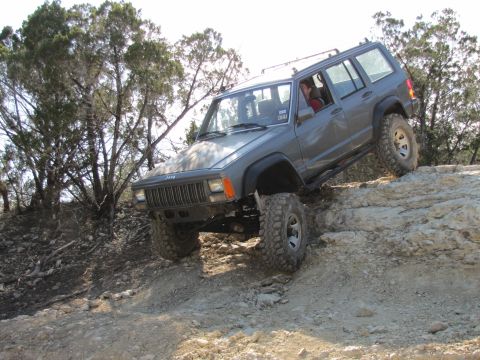 1995 Ford Bronco - A gray Jeep Cherokee is shown driving up a steep, rocky incline with trees in the background. The vehicle is angled towards the right, with…