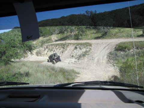 1995 Ford Bronco - A lone off-road vehicle navigates a winding dirt path surrounded by trees and hills.
