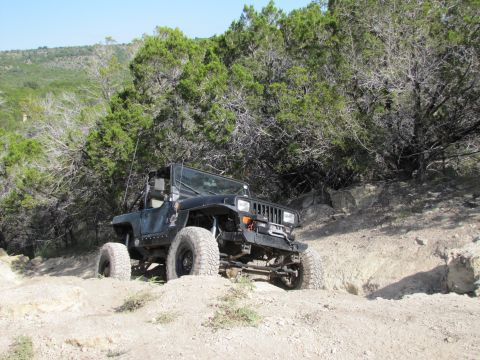 1995 Ford Bronco - A black Jeep Wrangler navigates a dirt path surrounded by trees and rocky terrain.