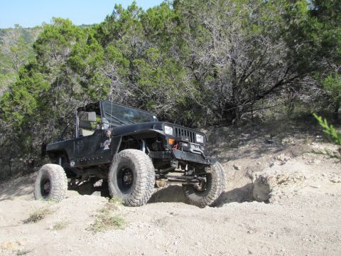 1995 Ford Bronco - Modified Jeep Wrangler navigating an off-road trail with rocky terrain and dense foliage.